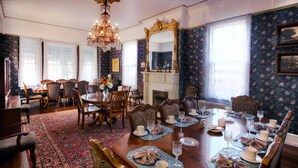 Un comedor formal con una gran mesa de madera dispuesta para una comida, rodeado de sillas ornamentadas, un candelabro y una chimenea.