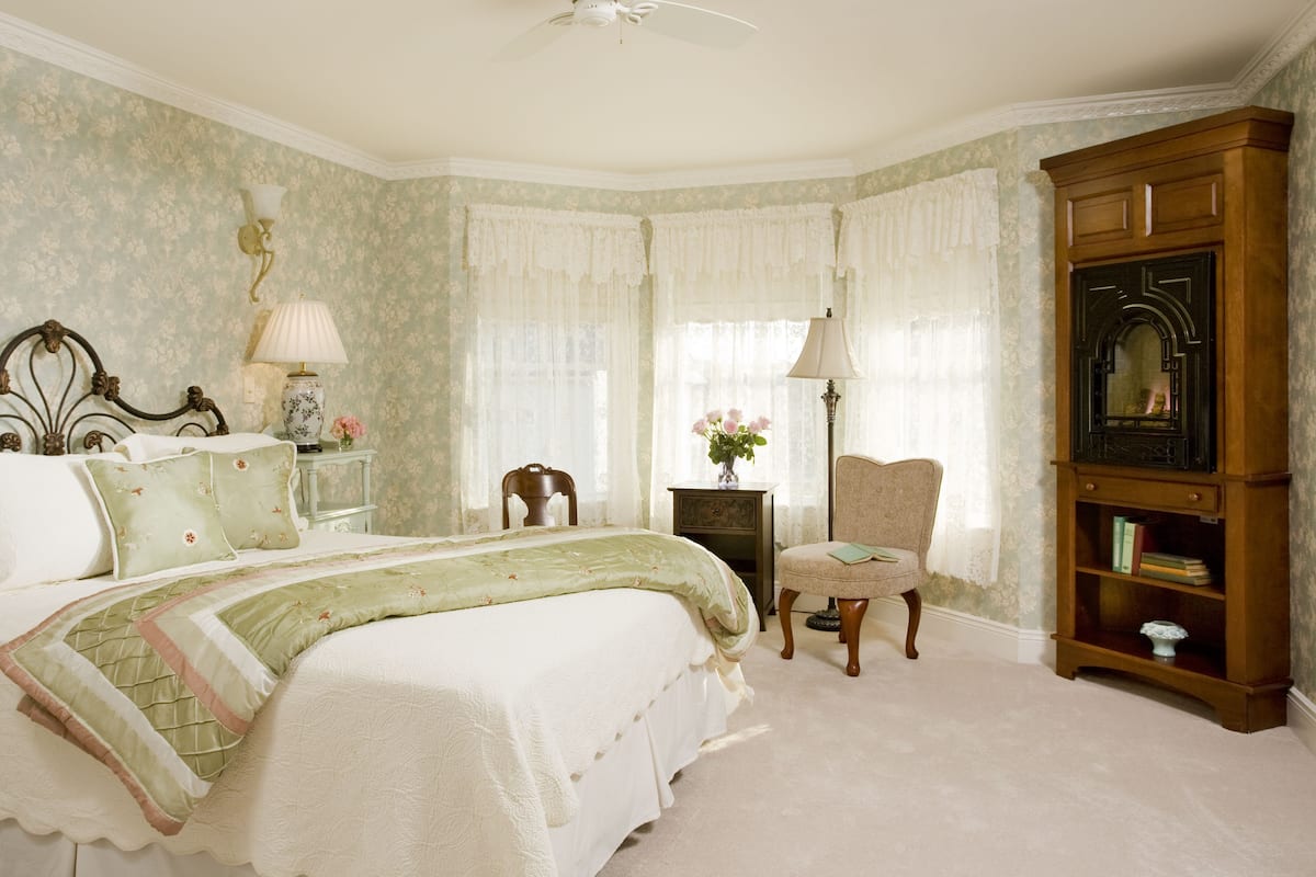 A bedroom with a bed, a chair, a bookshelf, and a nightstand with a lamp.