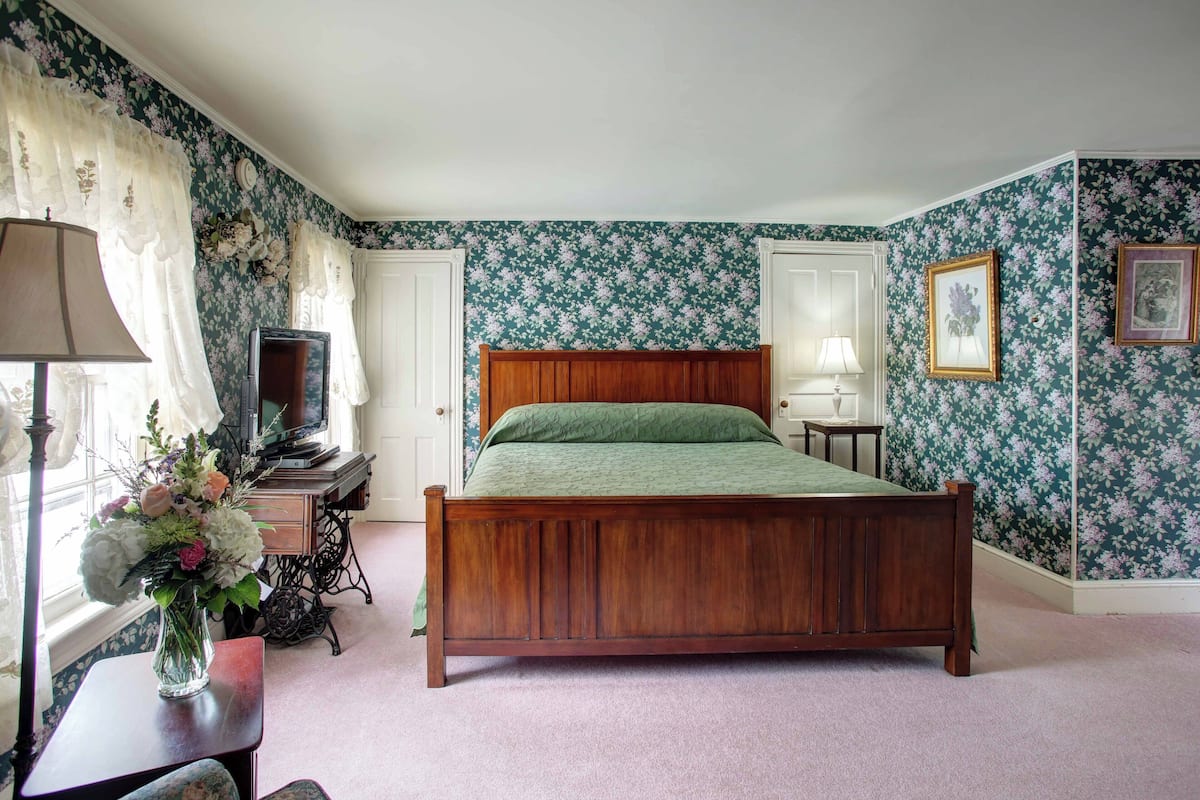 A bedroom with a wooden bed, a desk with a lamp, a television, and floral wallpaper.
