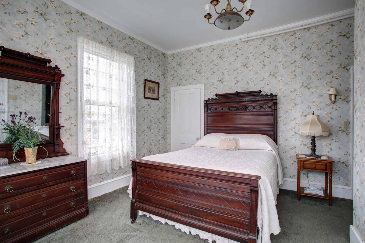 A bedroom with a wooden bed, a dresser, a mirror, a lamp, and floral wallpaper.