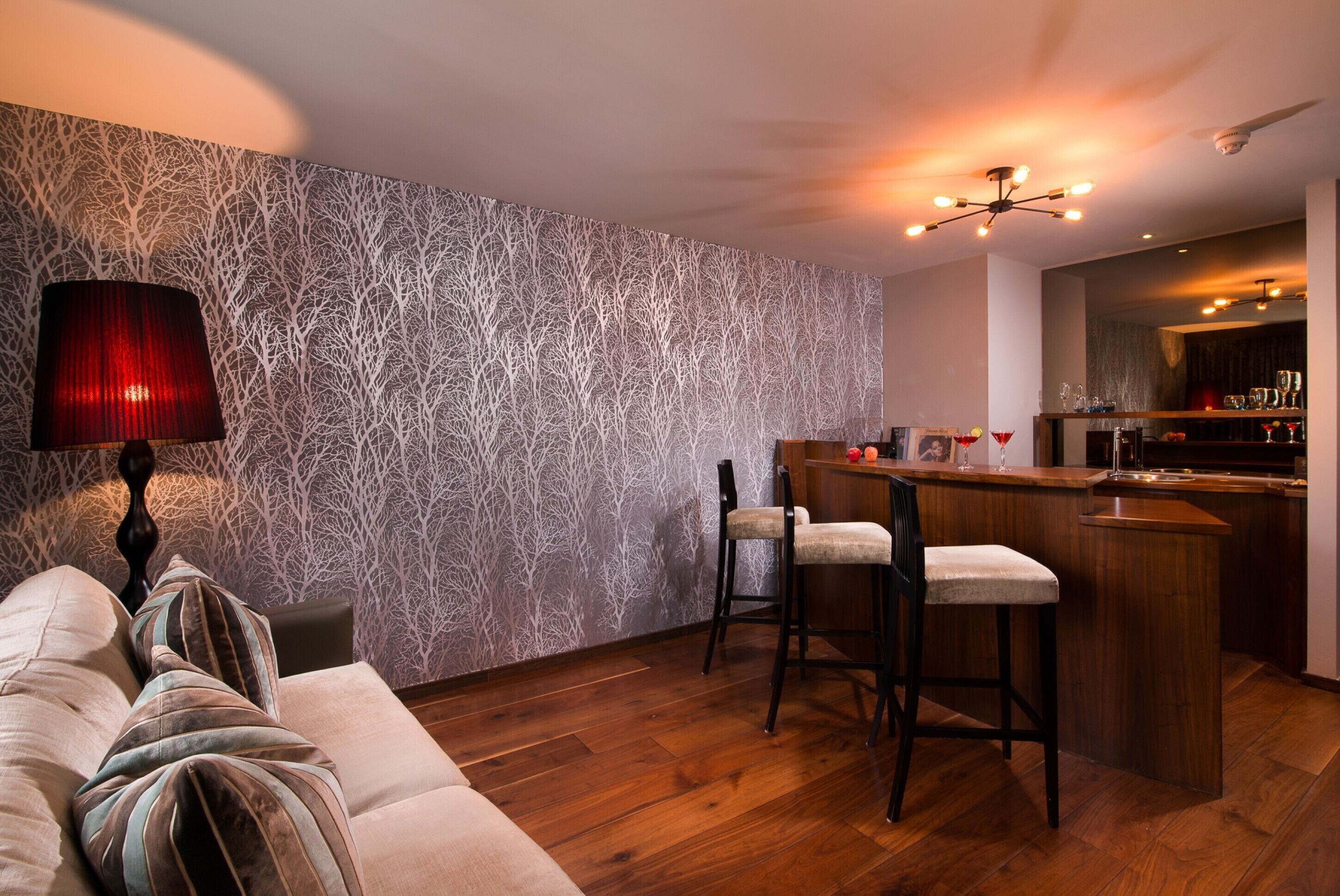 A modern living room with a patterned wallpaper, a sofa with striped pillows, a bar area with stools, and a ceiling fan.
