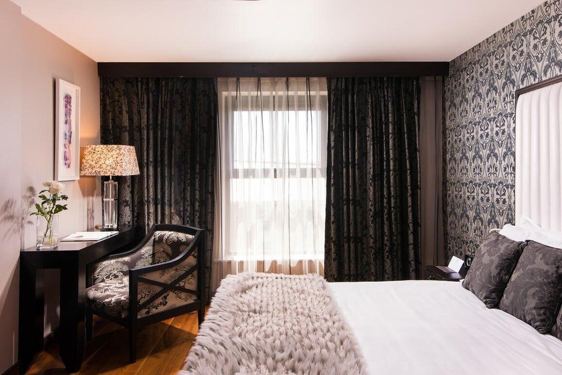 Une chambre d’hôtel avec un lit, un bureau avec une chaise, une lampe et une fenêtre avec des rideaux.