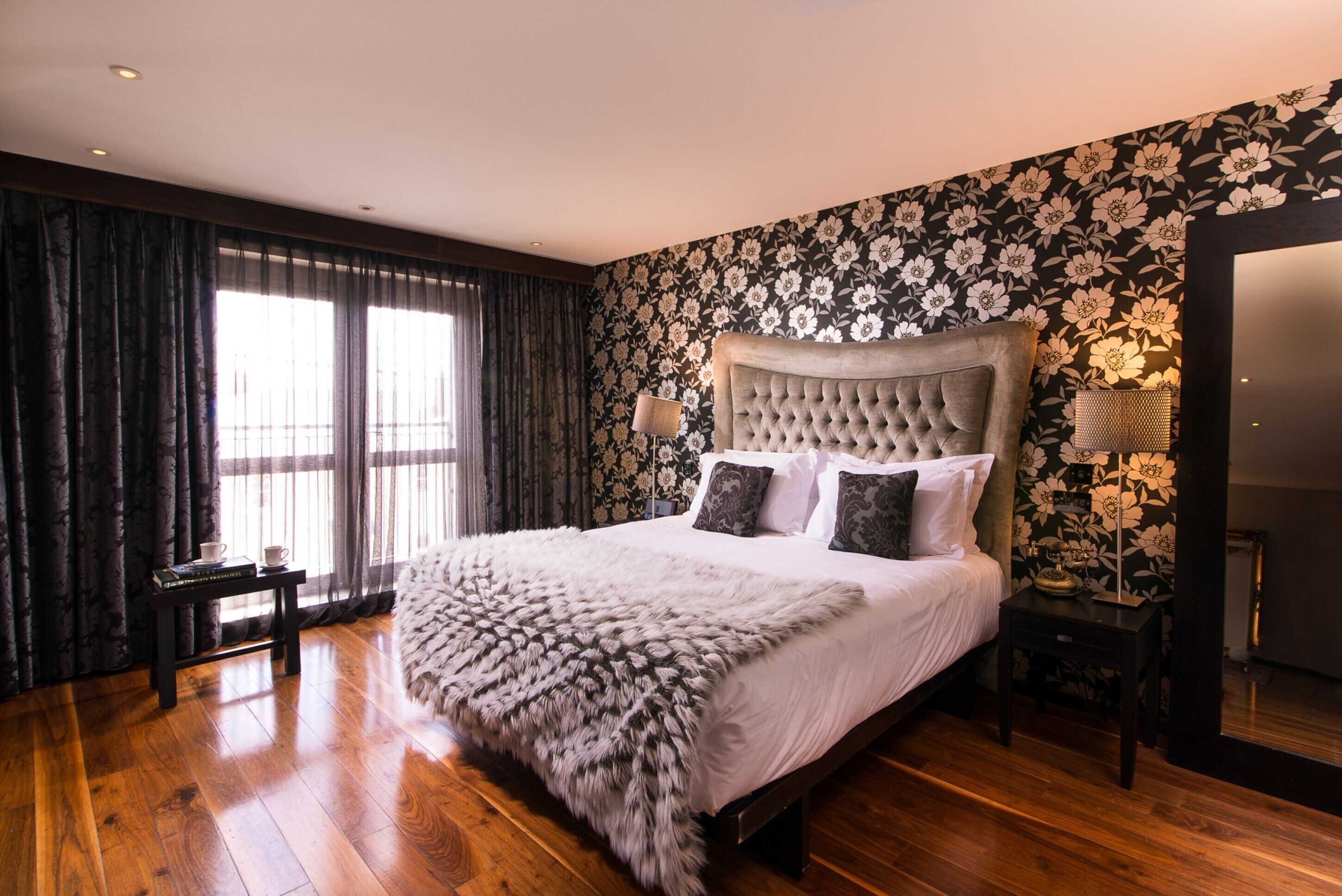 Quarto com uma cama grande, cortinas escuras, papel de parede floral e piso de madeira.