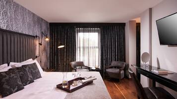 Habitación de hotel con una cama grande, dos sillones, un escritorio con televisor y una ventana con cortinas.