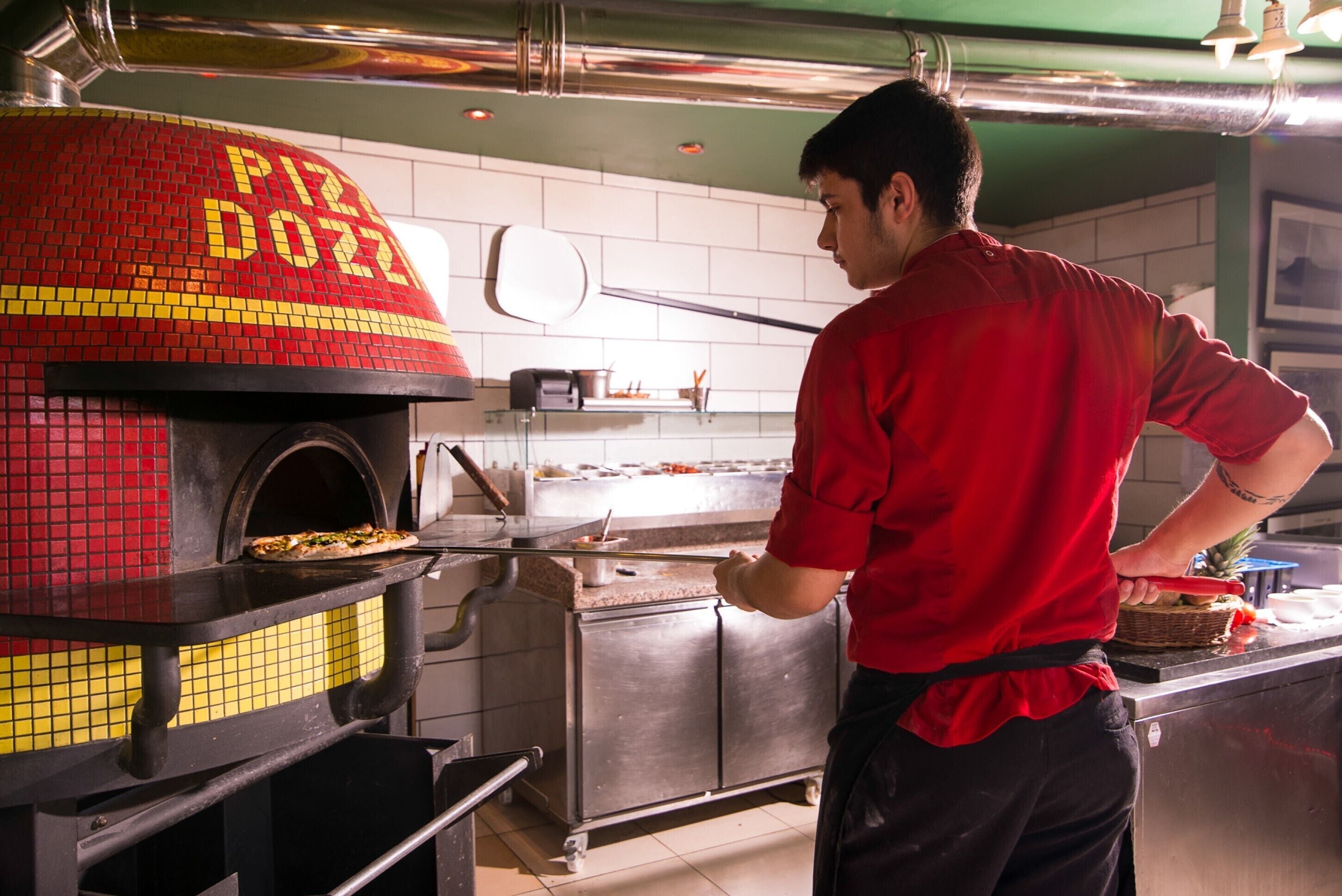 Una cocina con horno de pizza y una persona con camisa roja preparando comida.