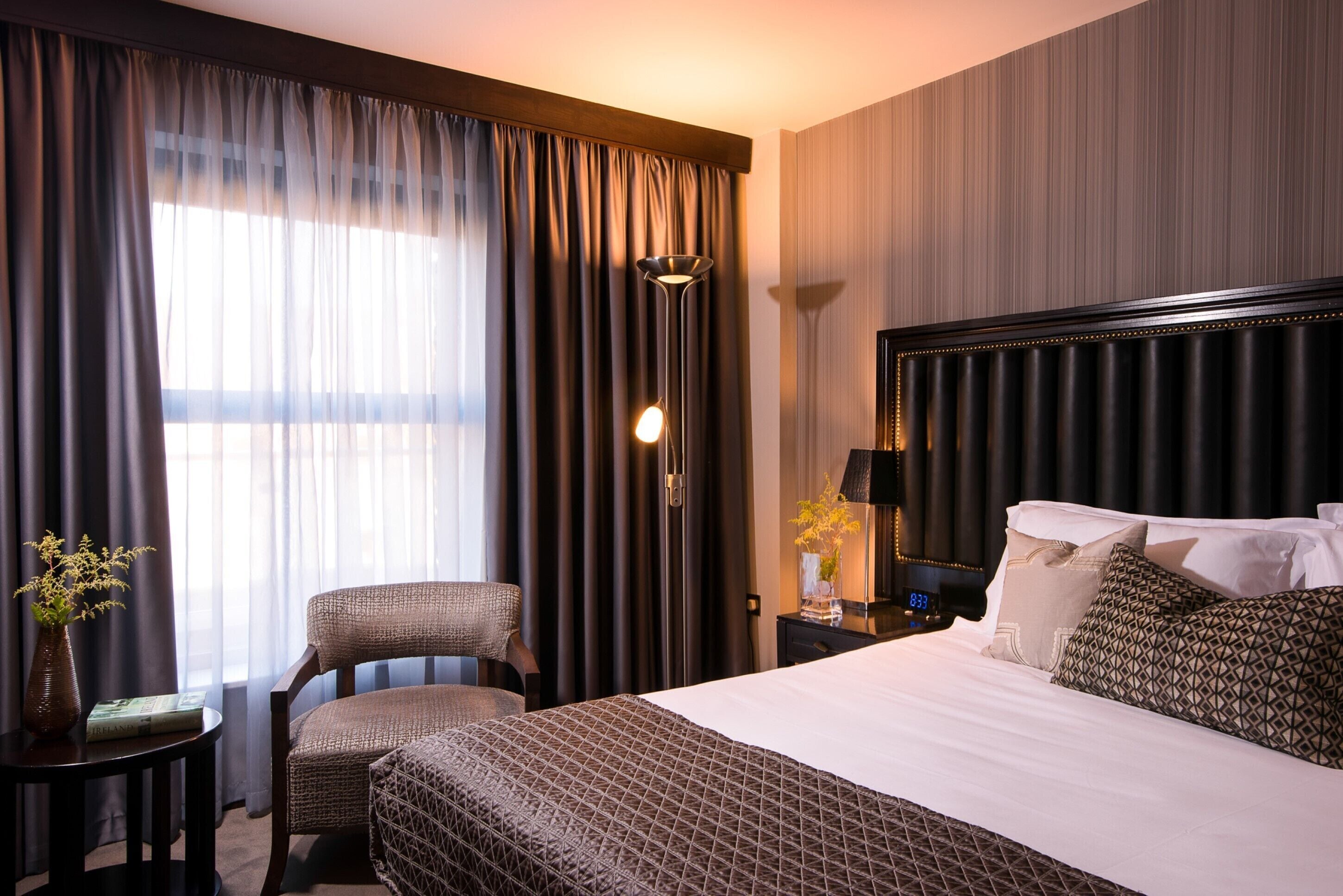 Habitación de hotel con una cama grande, una silla, una mesita de noche, una lámpara y una ventana con cortinas.