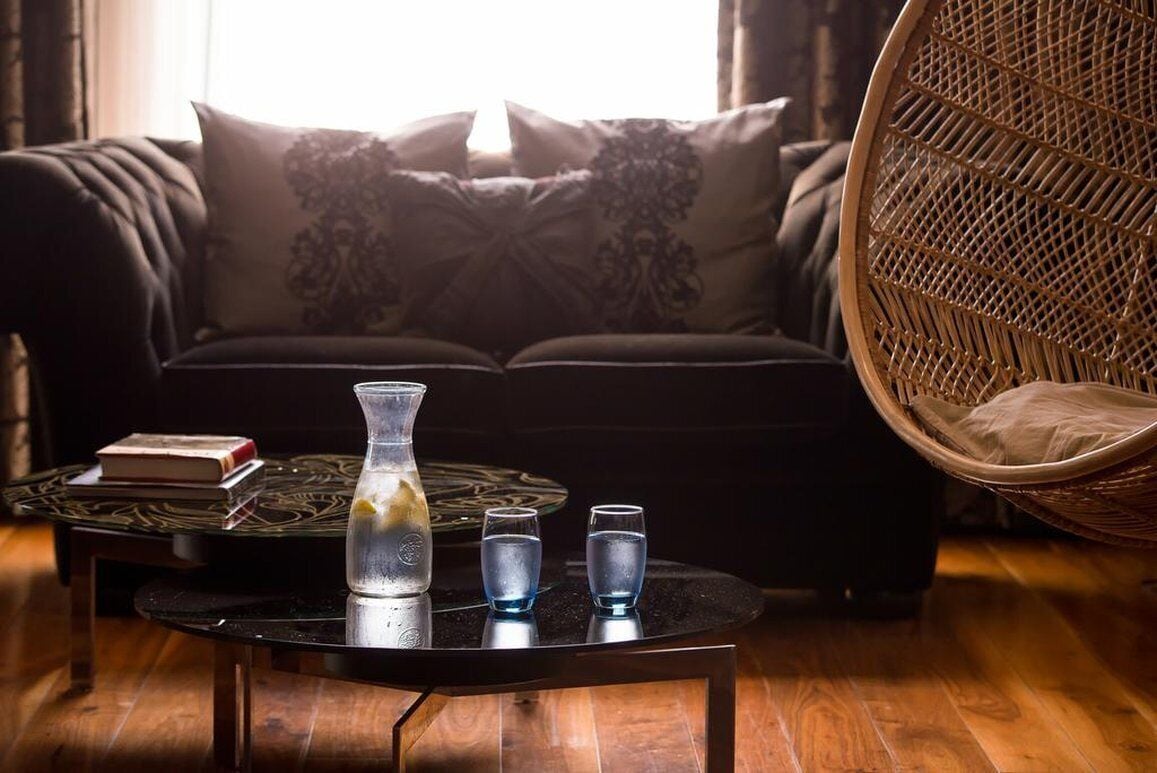 A cozy living room with a dark sofa, a wicker chair, a round coffee table with a vase, glasses, and a book.