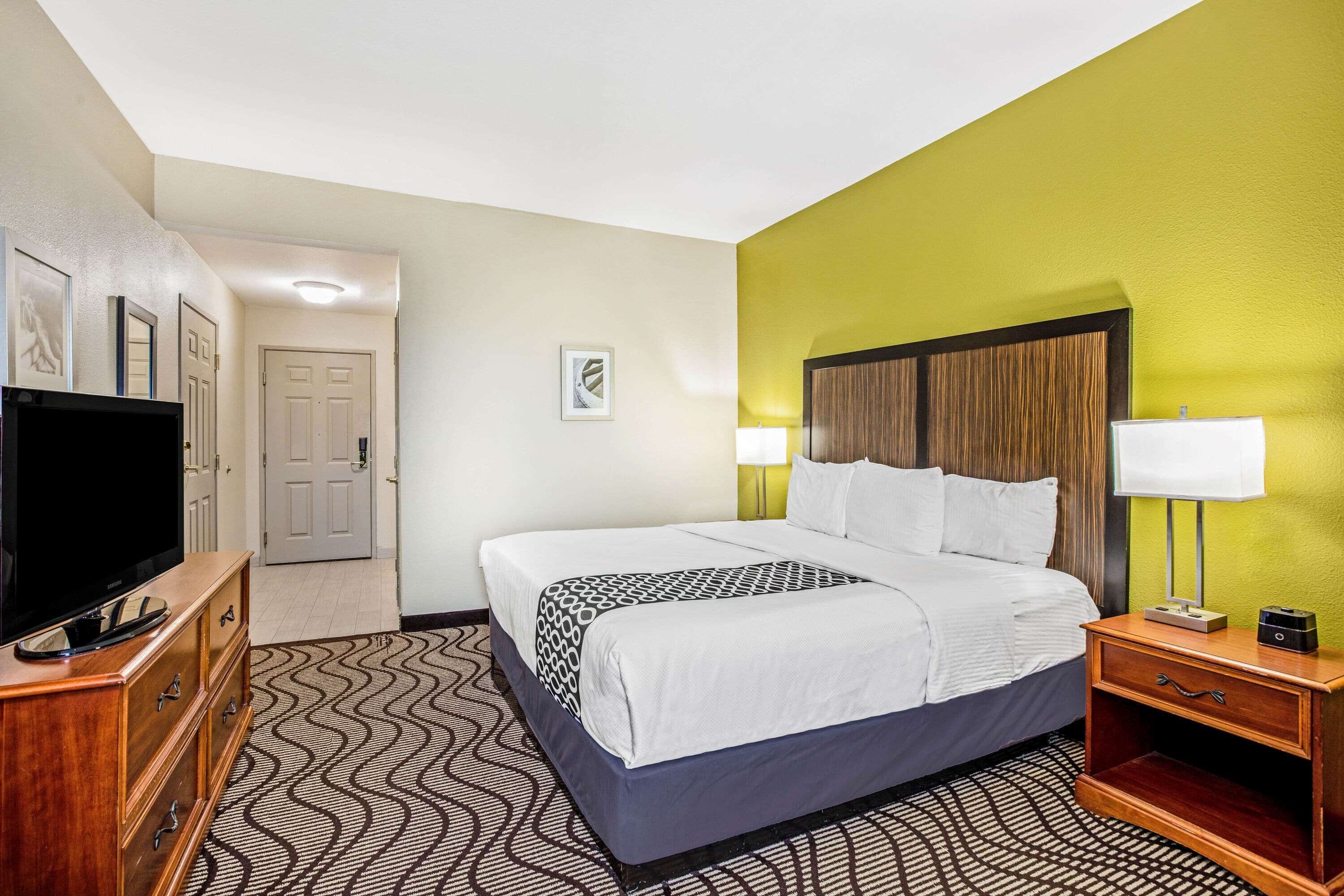 A hotel room with a large bed, a TV on a wooden dresser, a bedside table with a lamp, and a patterned rug.