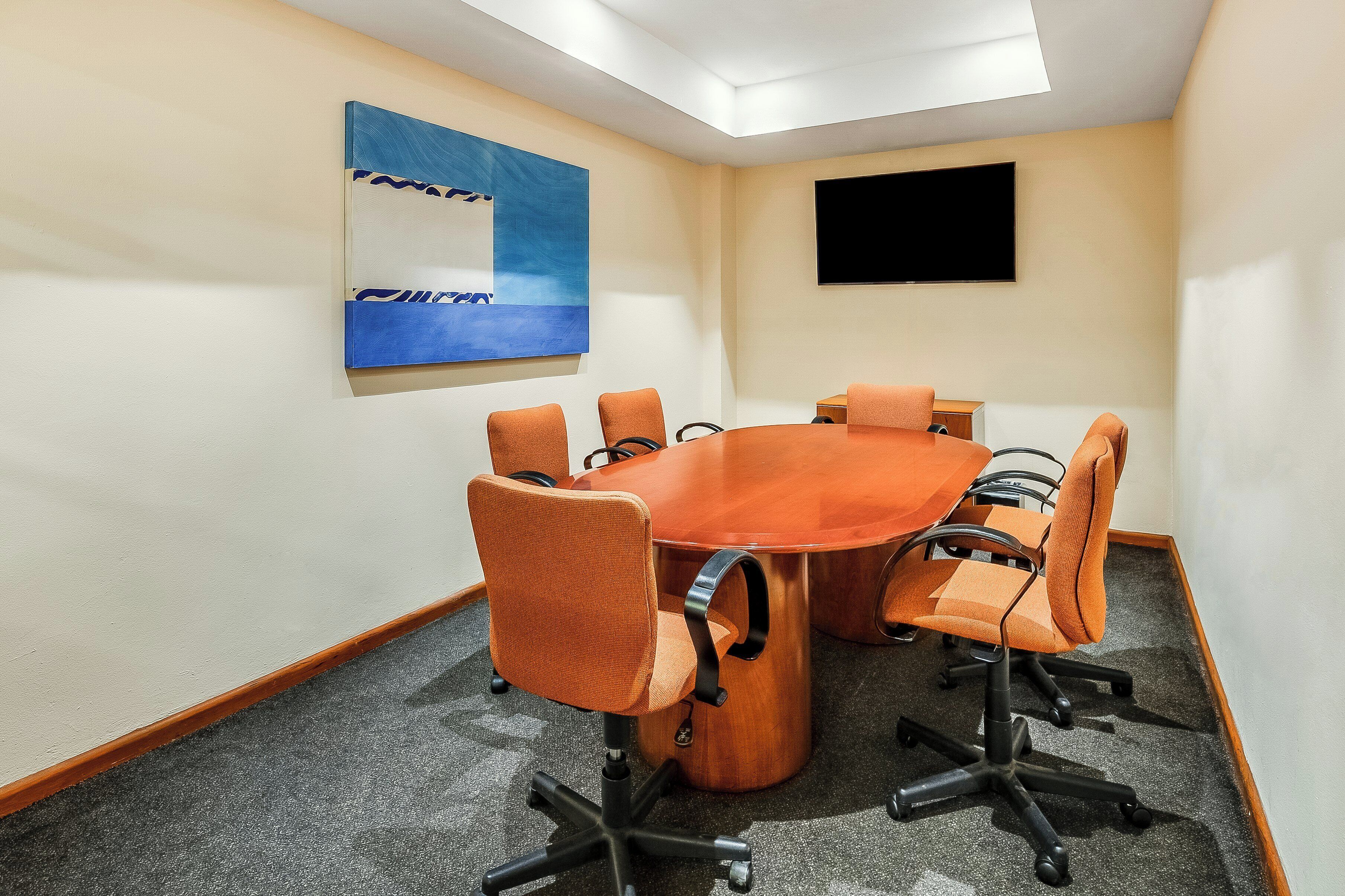 Un salón de conferencias con una mesa redonda, seis sillas anaranjadas, un cuadro abstracto grande y un televisor de pantalla plana en la pared.