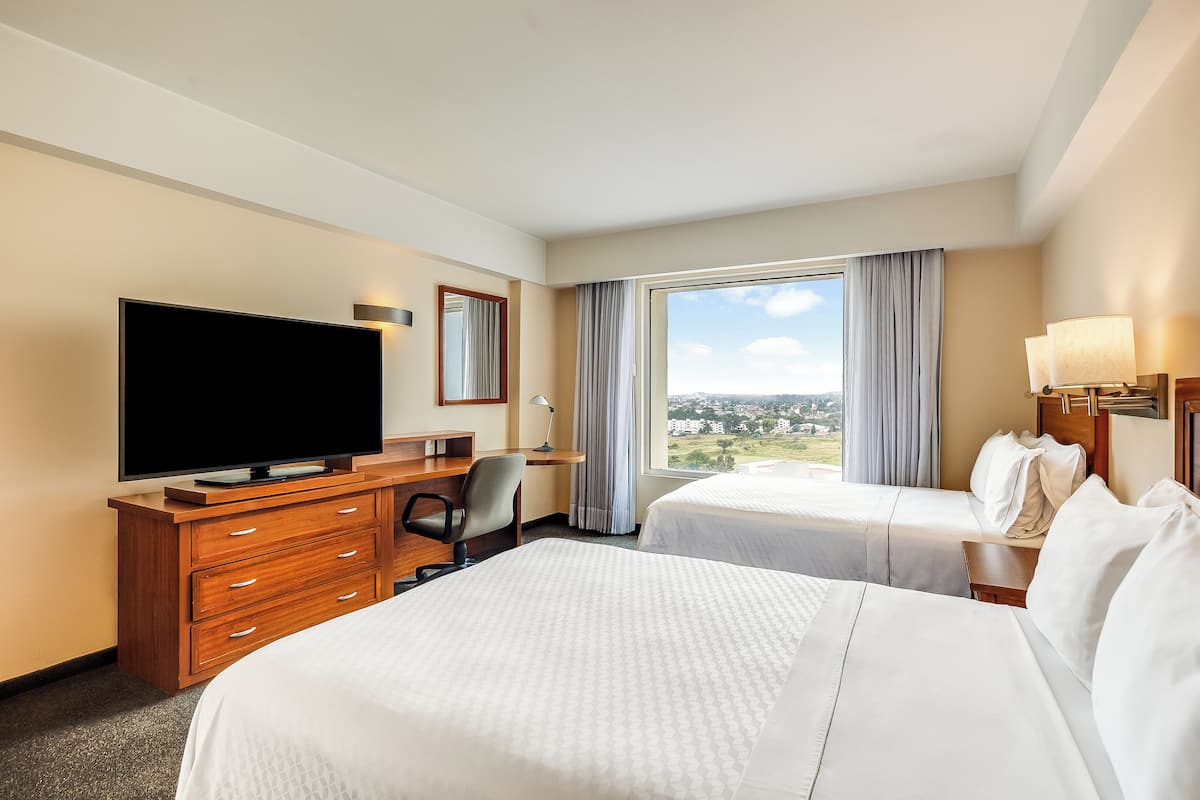 A hotel room with two beds, a TV, a desk, and a large window with a view.