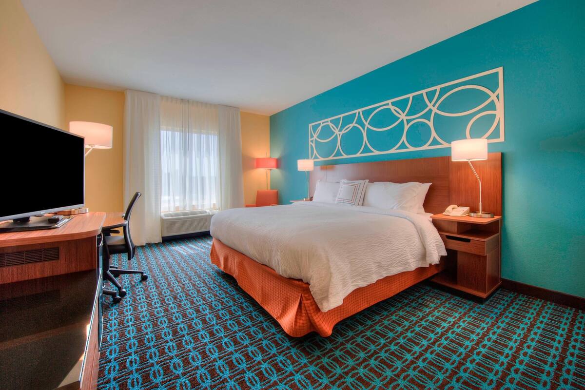 Ein Hotelzimmer mit Bett, Schreibtisch mit Fernseher, Sessel, Nachttisch mit Lampe und einem Fenster mit Vorhängen.