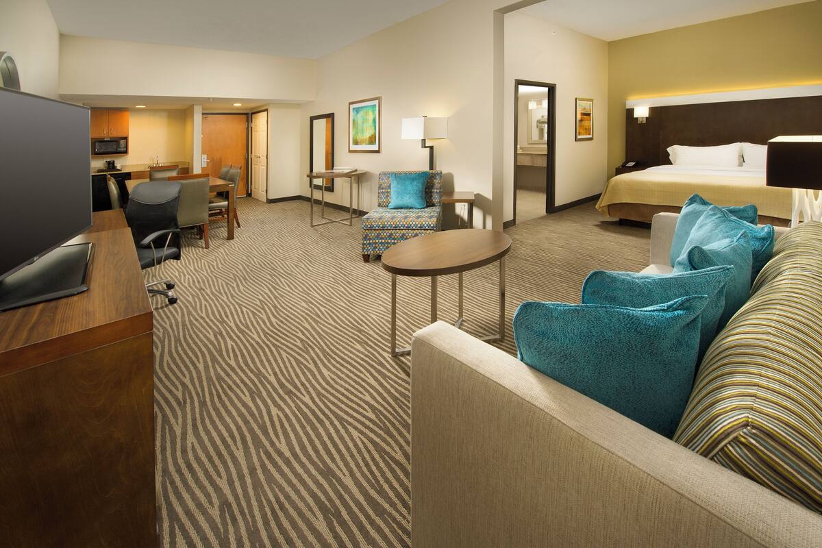 Habitación de hotel con cama, zona de estar con sofá y sillones, mesa de comedor con sillas, televisor y pequeña cocina.