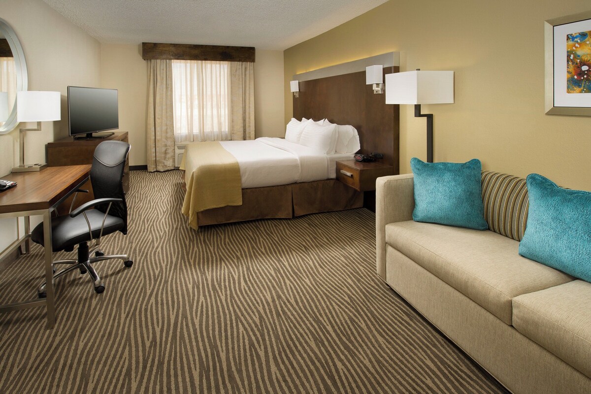A hotel room with a bed, a desk with a chair, a sofa, a television, and a painting on the wall.