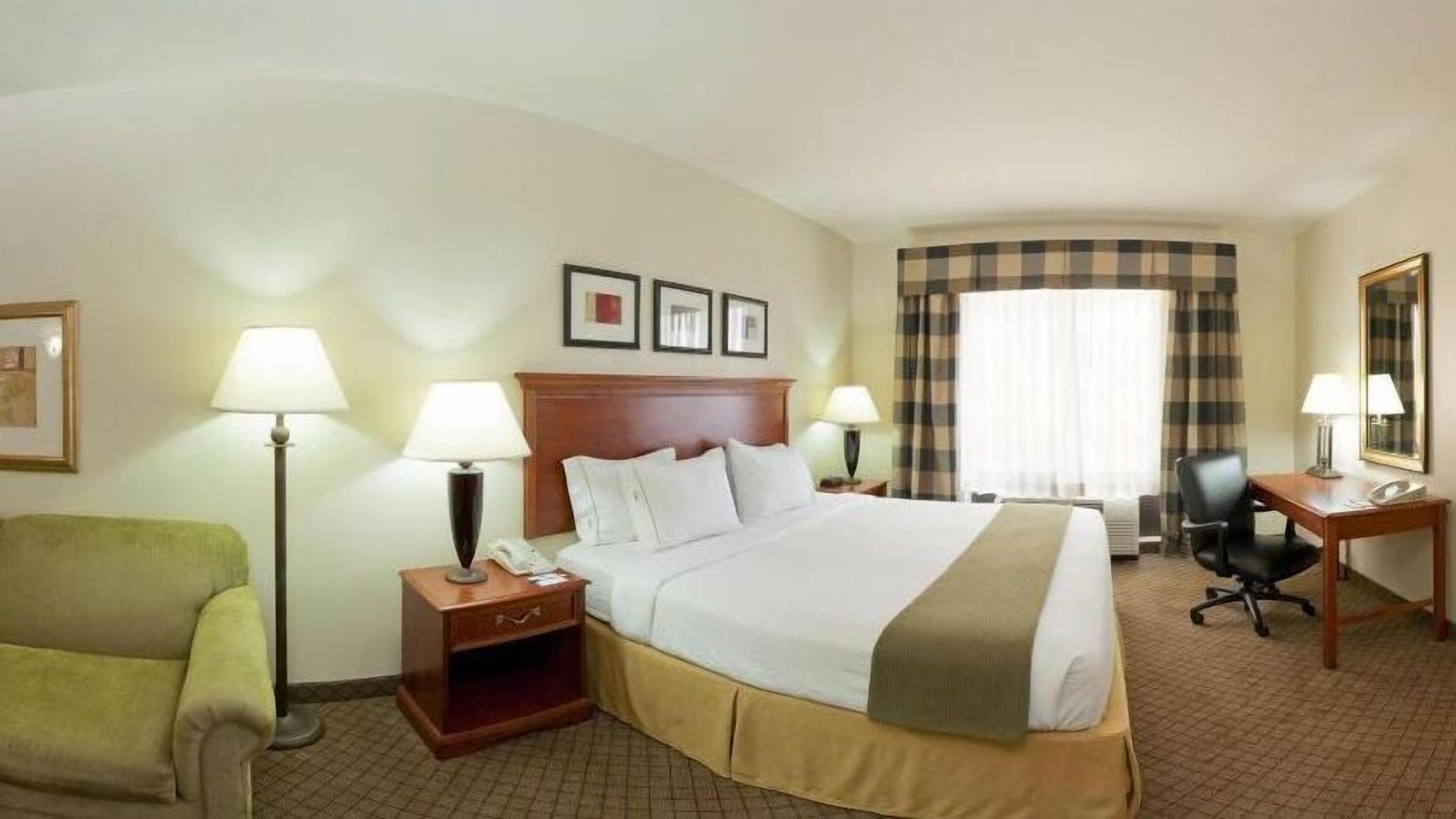 A hotel room with a large bed, two bedside tables with lamps, a green armchair, a desk with a chair, and a window with curtains.