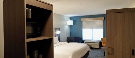 A hotel room with a bed, a wardrobe, a desk with a chair, a lamp, and a window with blinds.