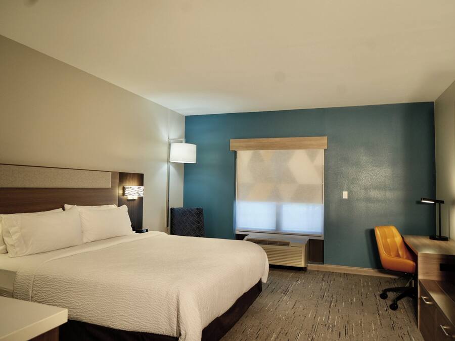 A hotel room with a large bed, a desk, an orange chair, a window with blinds, and a wall-mounted air conditioner.