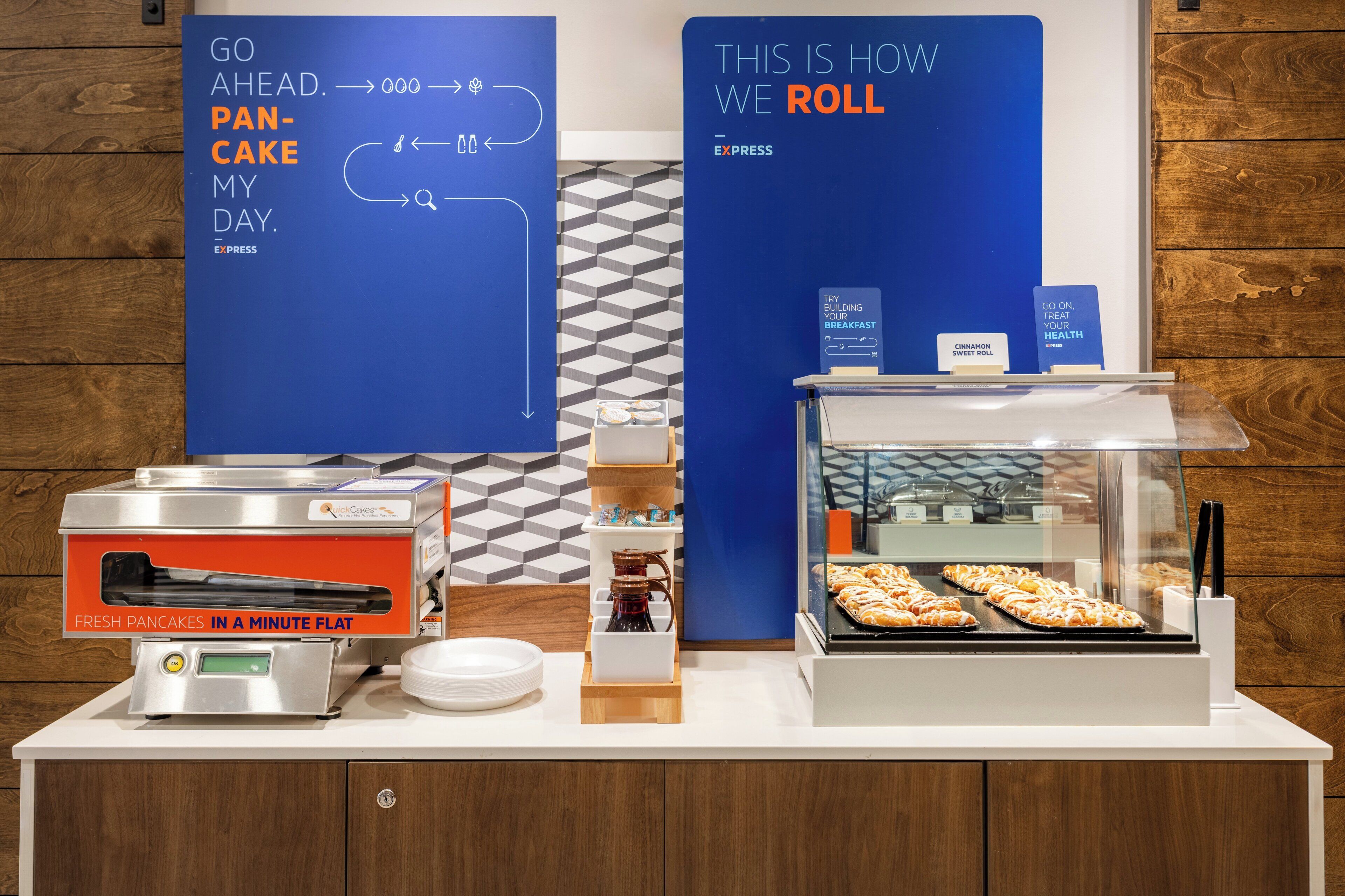 Una barra de desayuno con una máquina para hacer panqueques, una exhibición de pasteles e información en carteles.