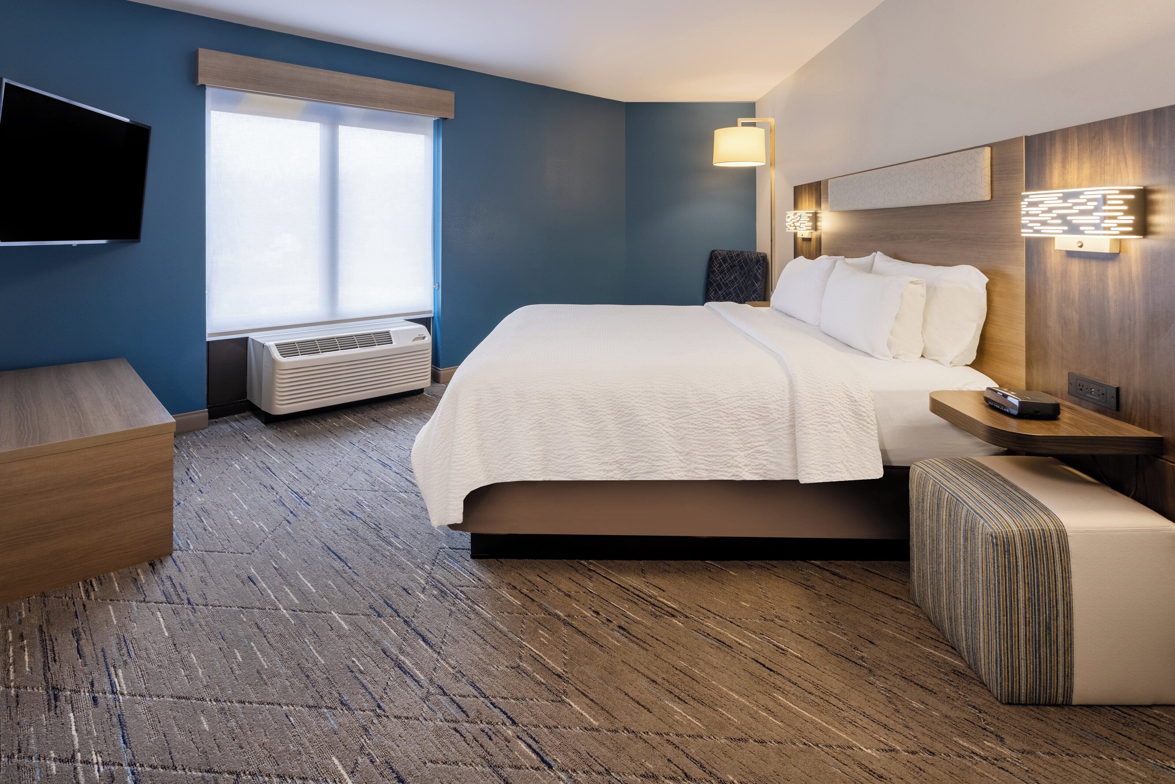 A modern hotel room with a large bed, a flat-screen TV, an air conditioning unit, and a wooden floor.
