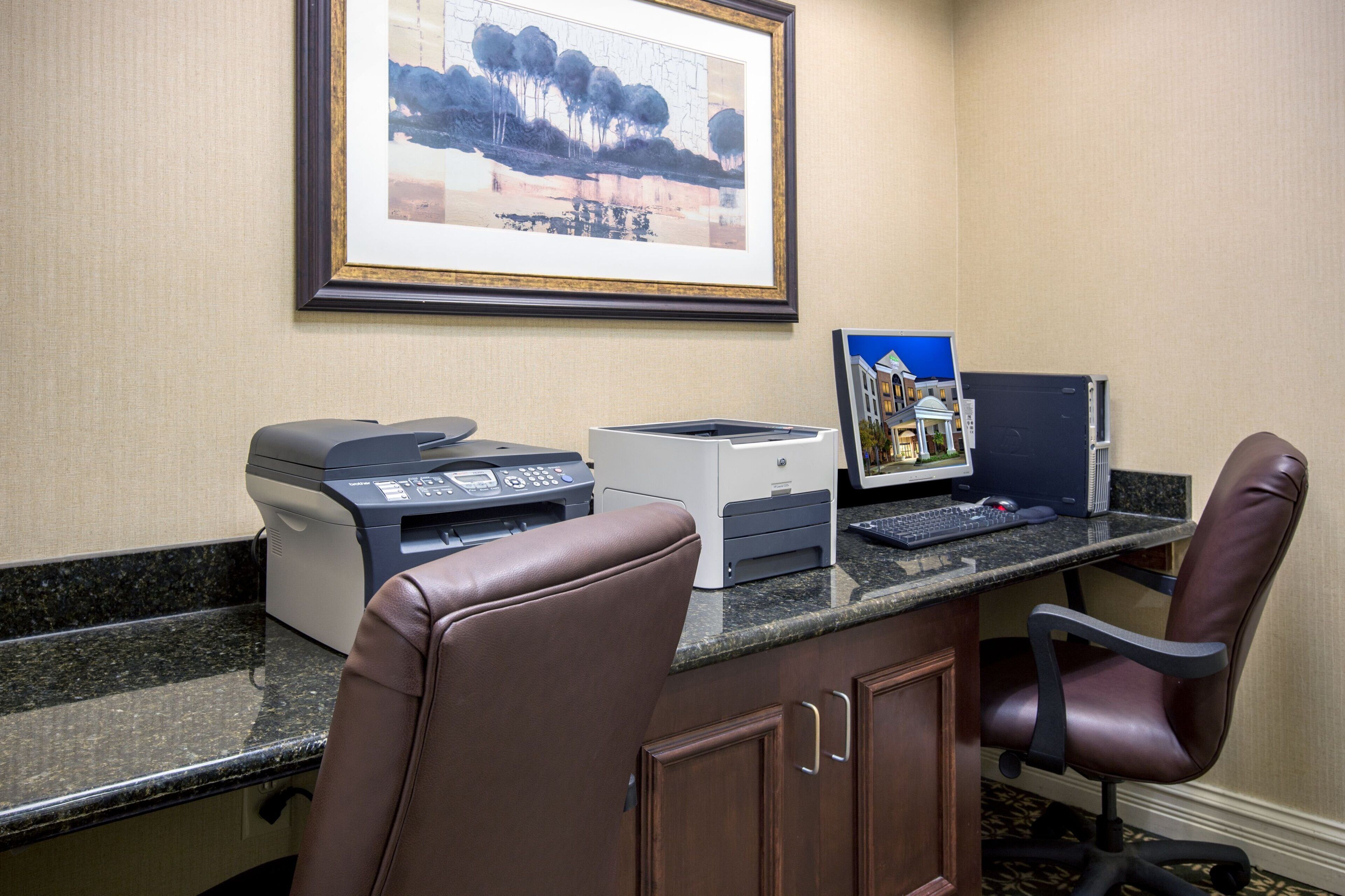 A hotel office with a desktop computer, a printer, and a framed landscape painting on the wall.