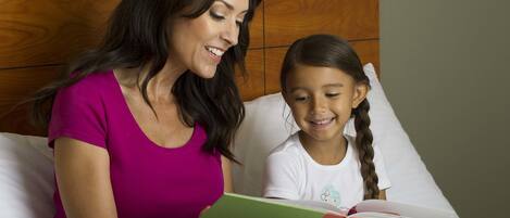 A woman and a child sitting on a bed with a wooden headboard, reading a book together.