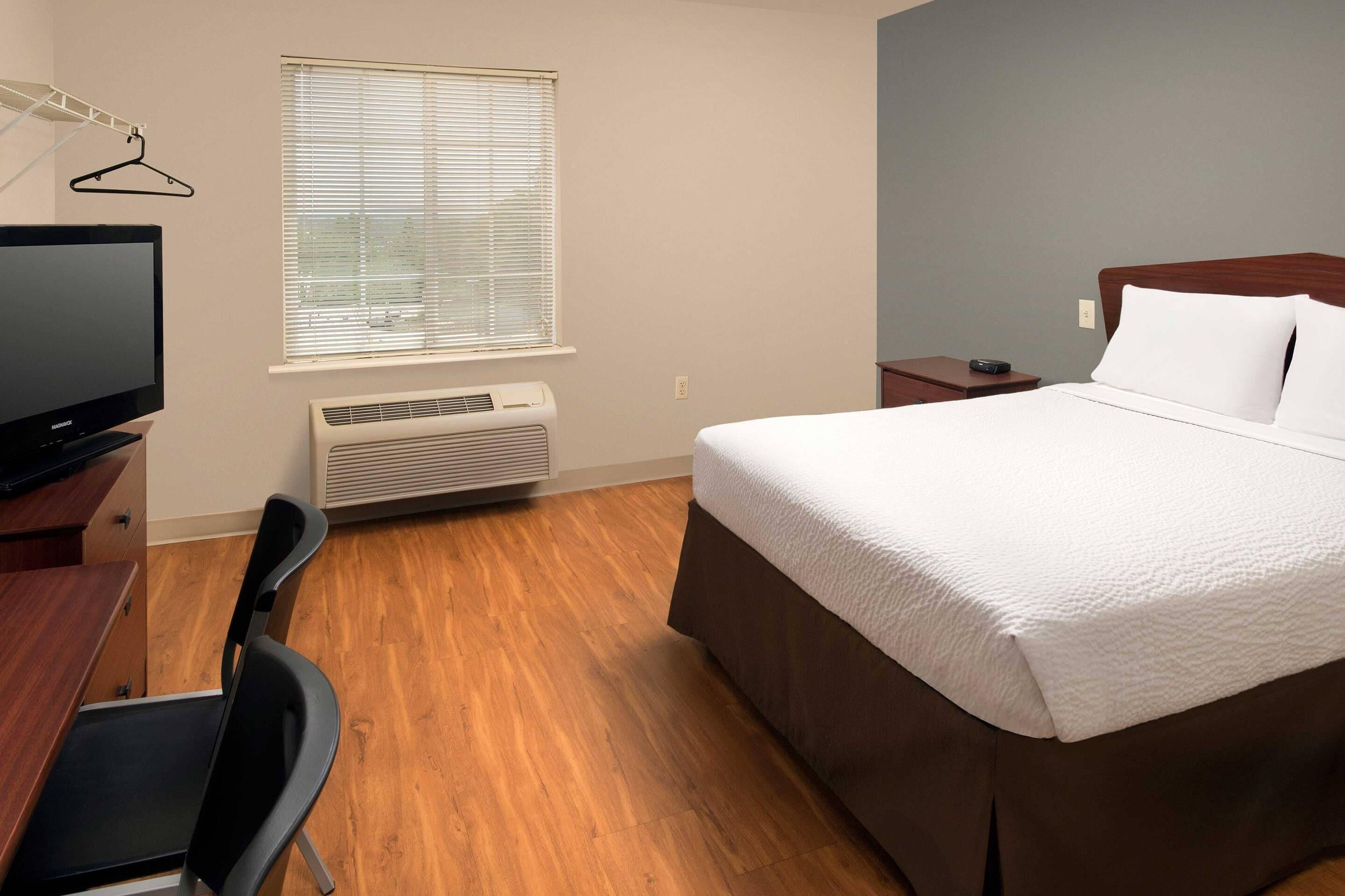 Een hotelkamer met een bed, een bureau met stoel, een televisie, een raam met jaloezieën en een airconditioner.