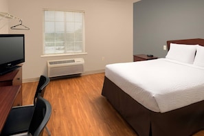 A hotel room with a bed, a desk with a chair, a television, a window with blinds, and an air conditioning unit. - WoodSpring Suites Kansas City Liberty (Liberty)