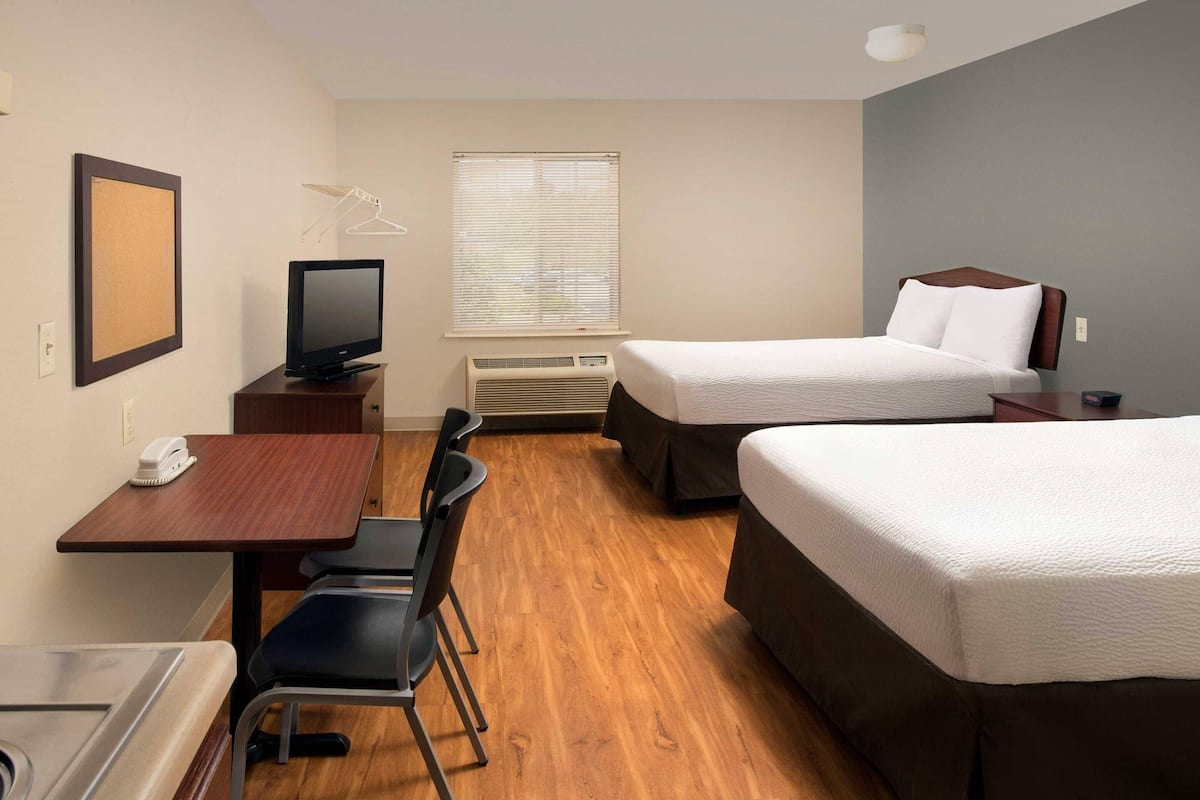 A hotel room with a desk, two beds, a television, a window with blinds, and a hanging clothes rack.