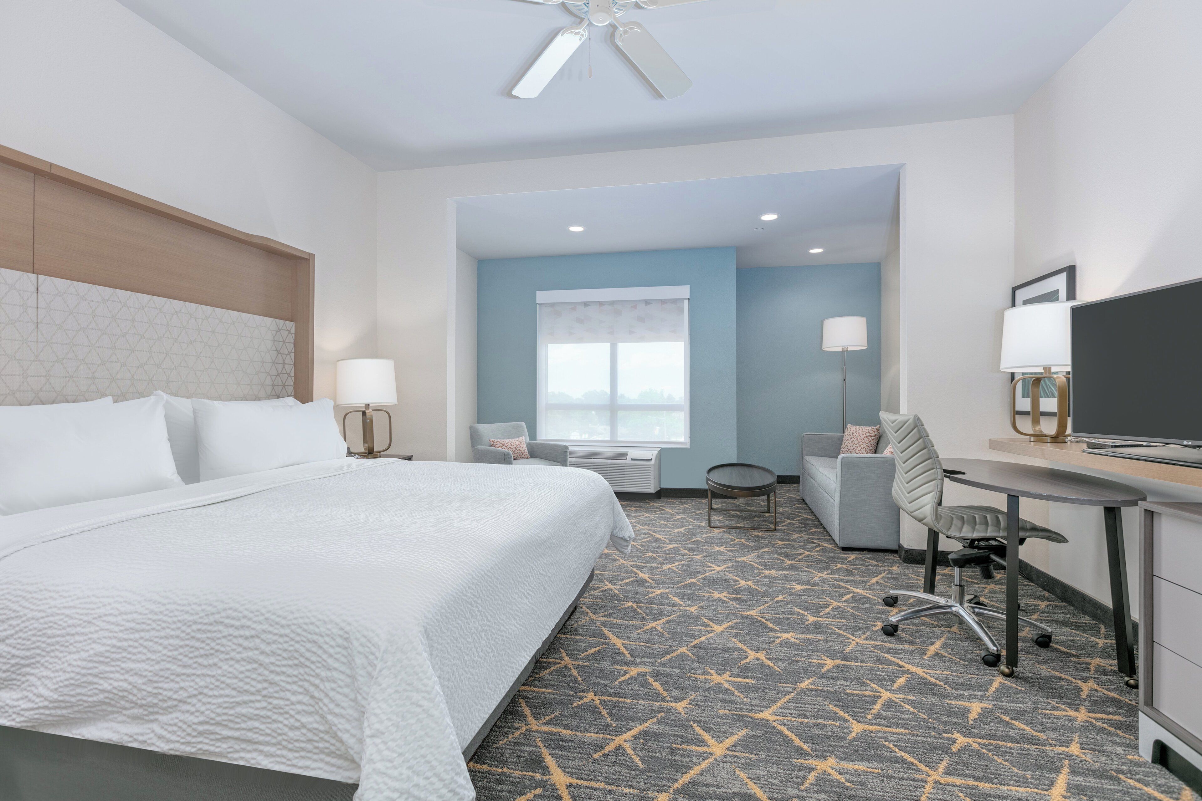 A hotel room with a large bed, a desk, a chair, a TV, and a ceiling fan.