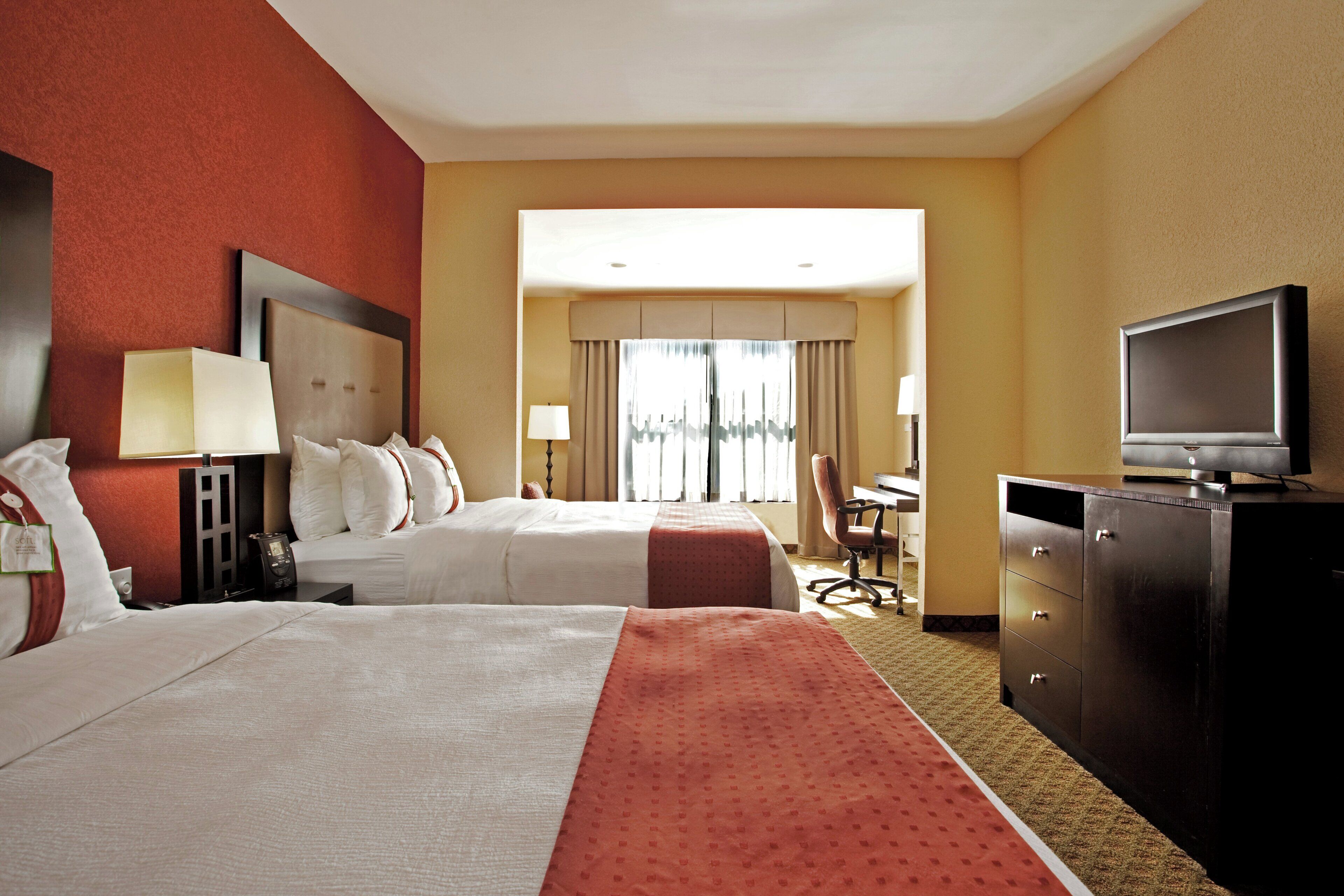 A hotel room with two beds, a desk, a television, and a view of the city.