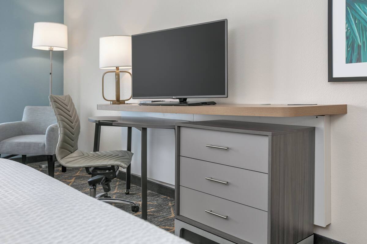 A hotel room with a bed, a desk with a television, a chair, a desk lamp, and a cabinet.