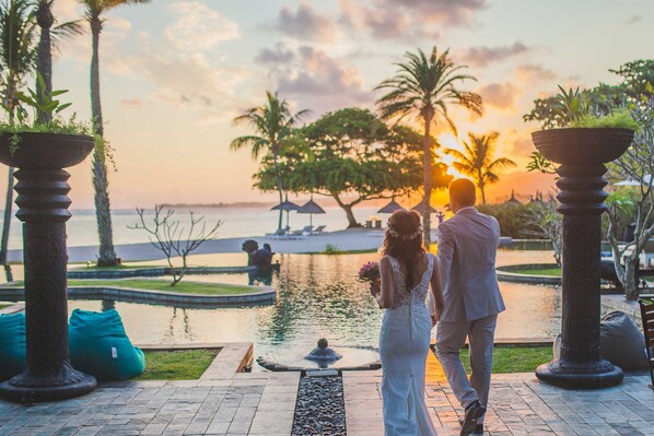Un couple se promène le long d’une promenade en bord de mer, avec en toile de fond un coucher de soleil, des palmiers et une petite fontaine.