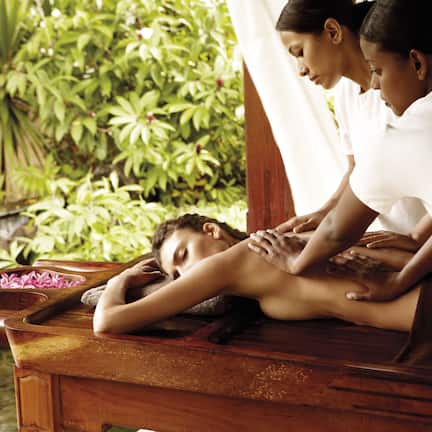 A spa setting with a wooden massage table, a person receiving a massage, and a lush green background.