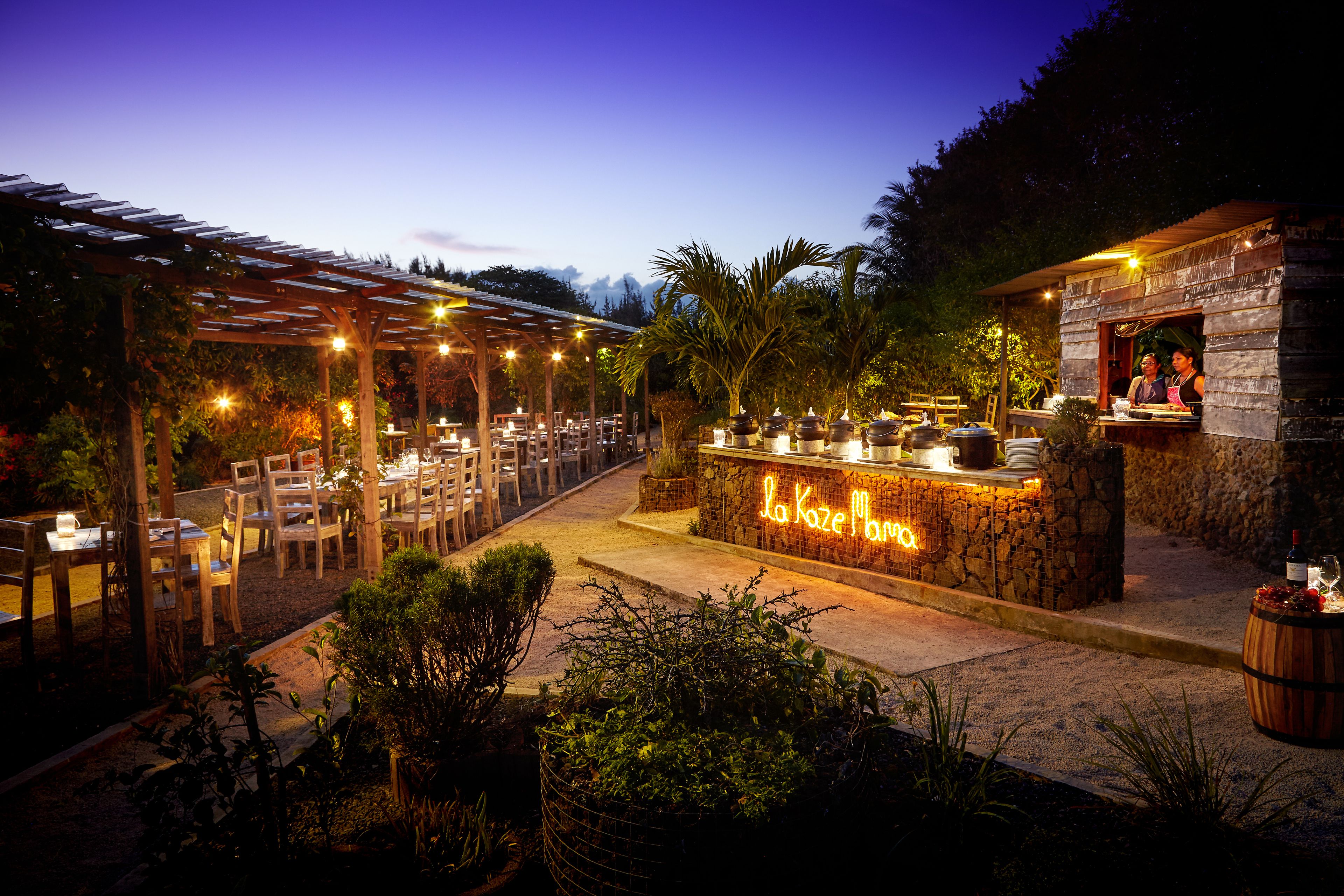 An outdoor restaurant with wooden tables and chairs, a stone bar, and a pergola with string lights.