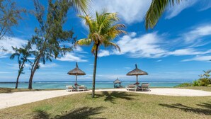 A beach with three thatched umbrellas, two lounge chairs, and a palm tree. - Shanti Maurice Resort & Spa (Chemin Grenier)