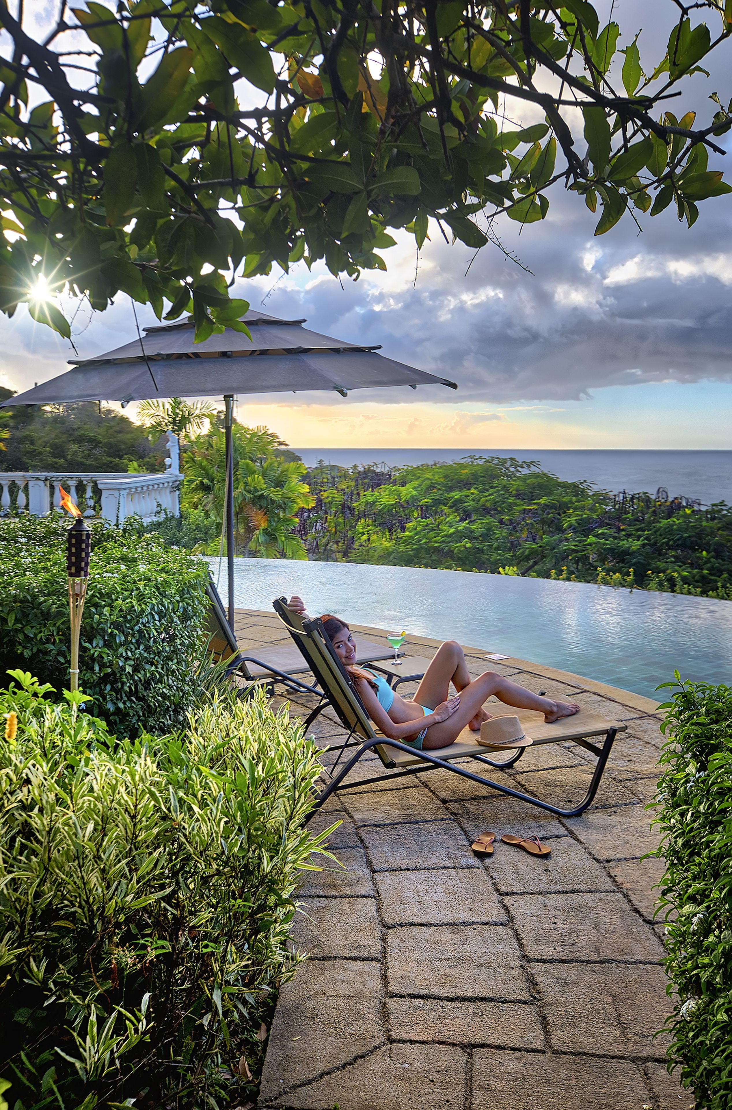 Une personne se détend sur une chaise longue au bord d’une piscine, avec vue sur l’océan et une végétation luxuriante.