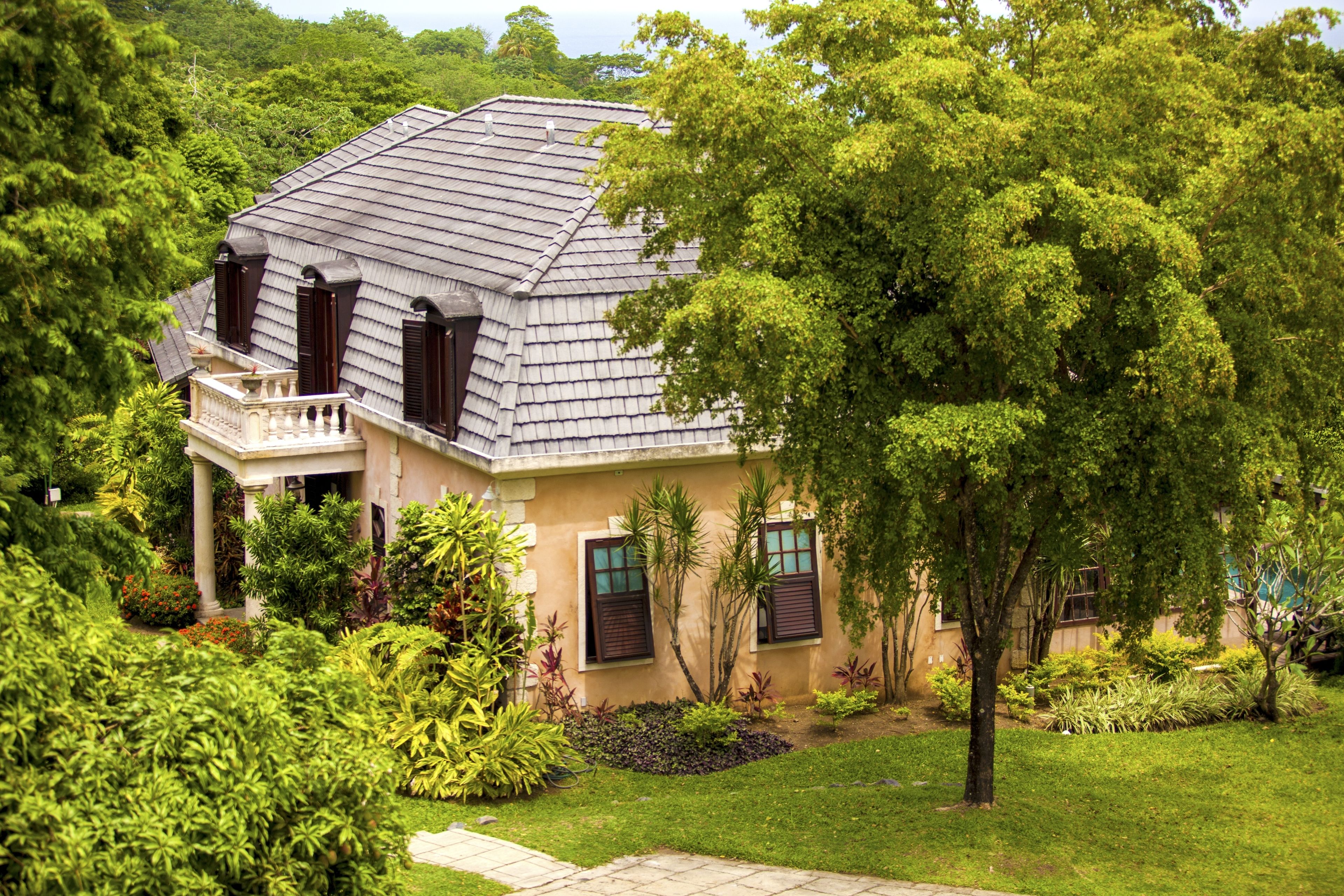 Uma casa de dois andares com telhado inclinado, varanda e um jardim bem cuidado com diversas plantas e gramado.