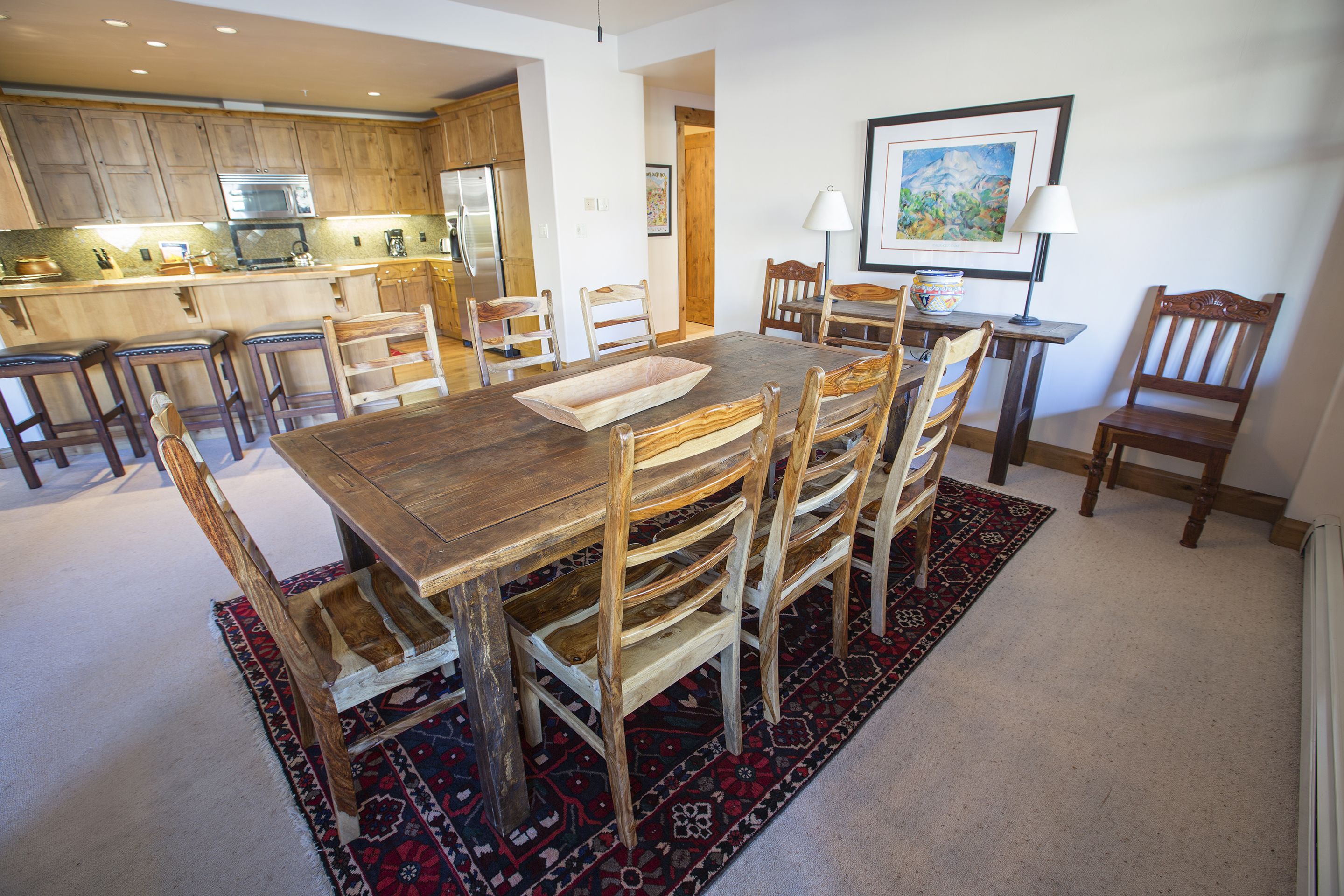 A dining area with a wooden table and chairs, a kitchen in the background, and a framed painting on the wall.