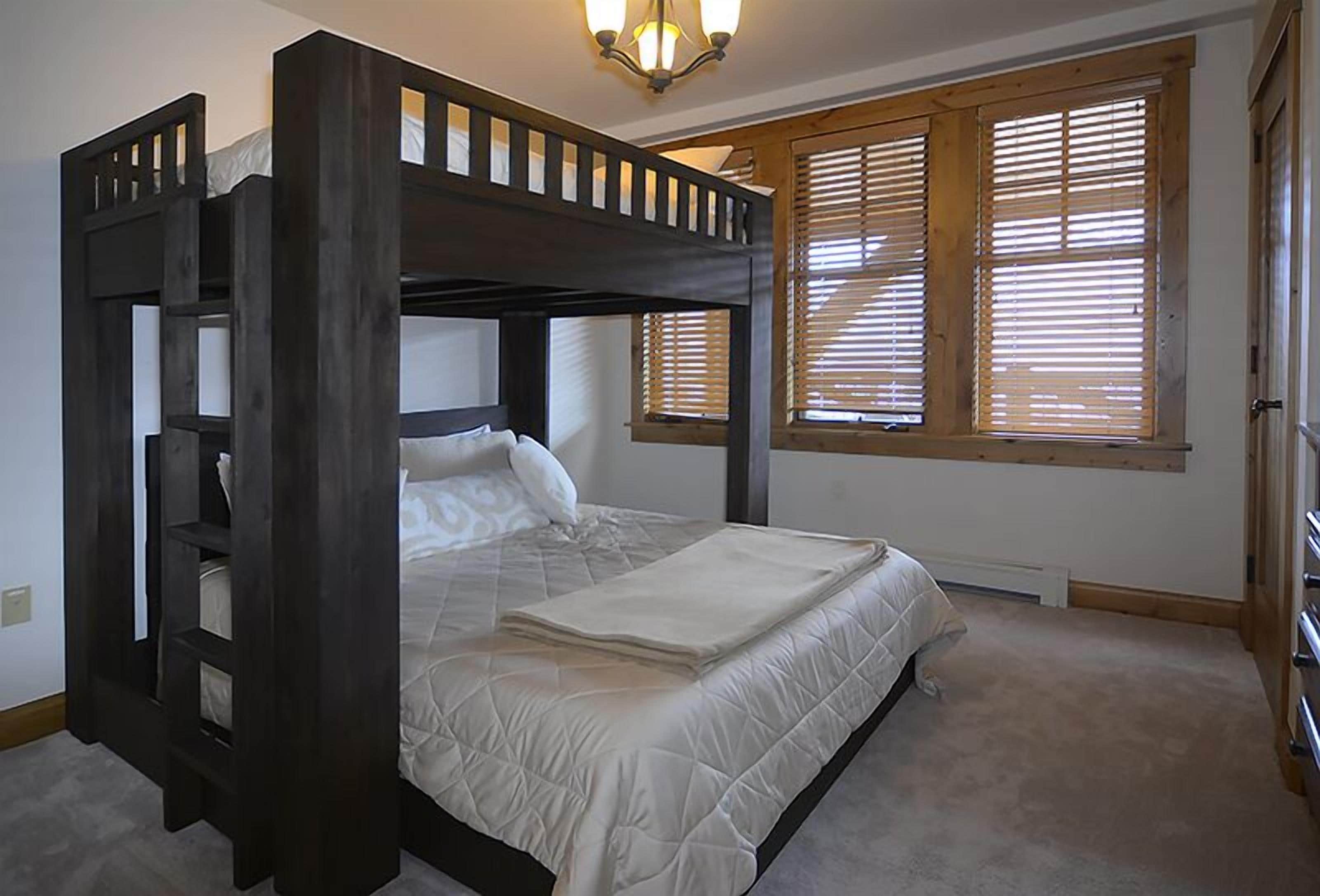 Una cama con dosel de madera y ropa de cama blanca en una habitación con alfombra color arena y persianas de madera en las ventanas.