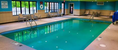 An indoor swimming pool with clear water, a lifeguard sign, and several chairs around the pool area.