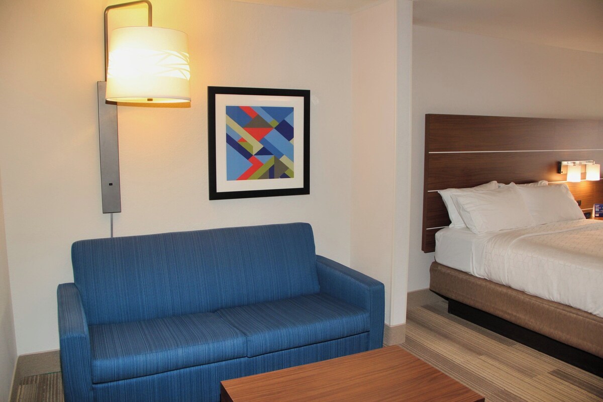 Una habitación de hotel con un sofá azul, una mesa de centro de madera, una cama con ropa blanca, una obra de arte abstracta enmarcada y una lámpara fijada a la pared.