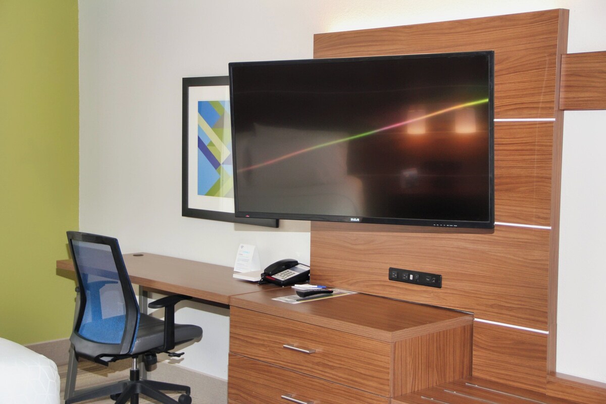 Una habitación de hotel moderna con un escritorio de madera, una silla de oficina negra, un televisor de pantalla plana y un cuadro en la pared.