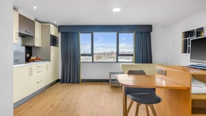 A modern kitchen with wooden cabinets, a dining table, and a view of the city through large windows. - Roomzzz Leeds City West (Leeds)