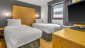 A hotel room with two beds, a wooden headboard, a large window with curtains, a flat-screen TV, and a wooden desk. - Roomzzz Leeds City West (Leeds)