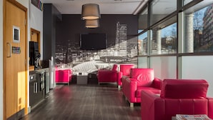 A modern hotel lobby with a cityscape view, red seating, a coffee machine, and a flat-screen TV. - Roomzzz Leeds City West (Leeds)
