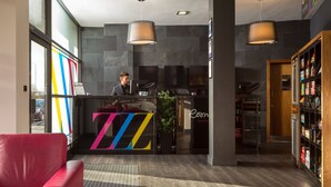 A modern hotel lobby with a reception desk, a red sofa, a large plant, and a wooden bookshelf. - Roomzzz Leeds City West (Leeds)