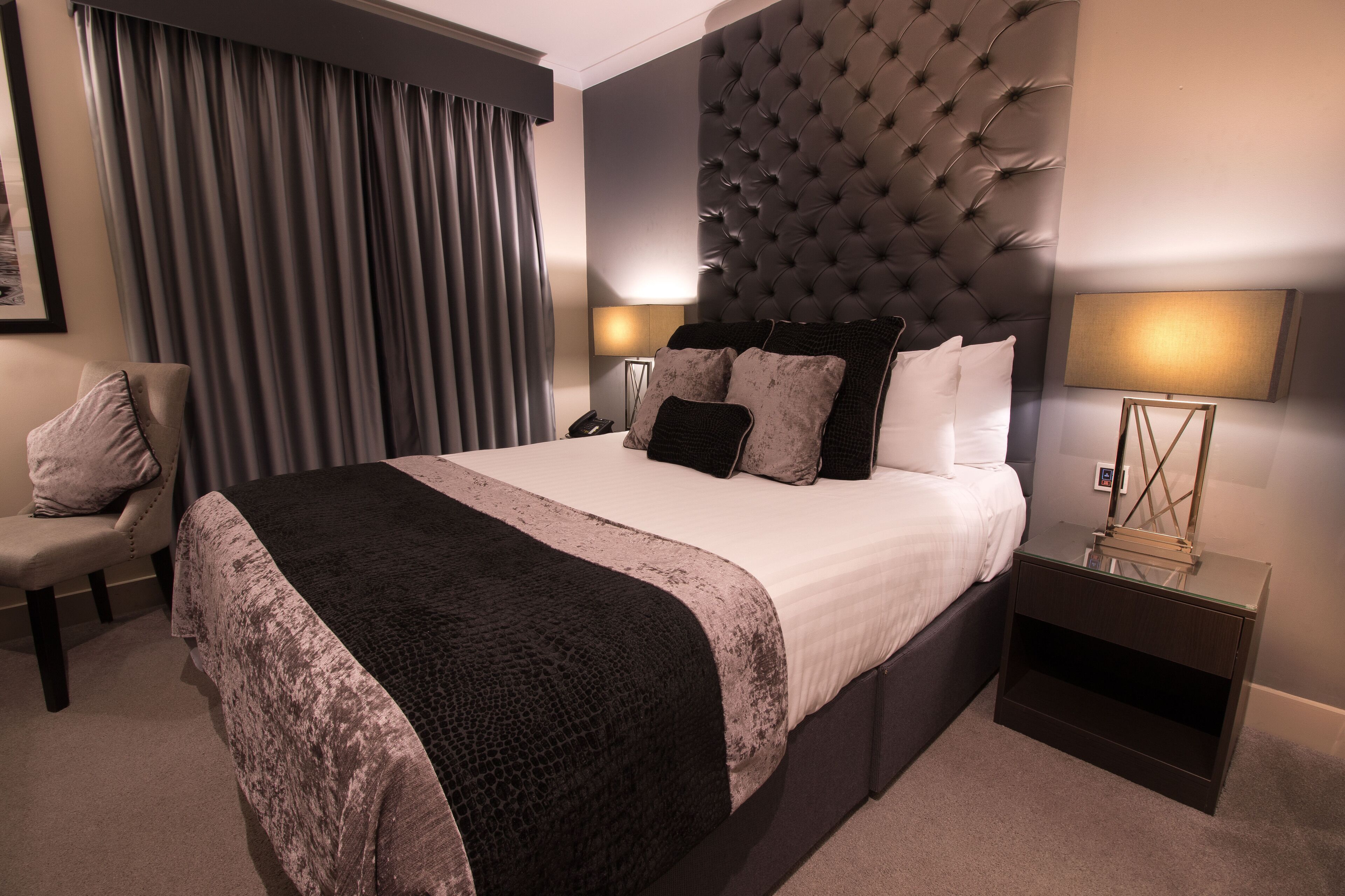 A modern hotel room with a large bed, a tufted headboard, a bedside table with a lamp, and a chair.