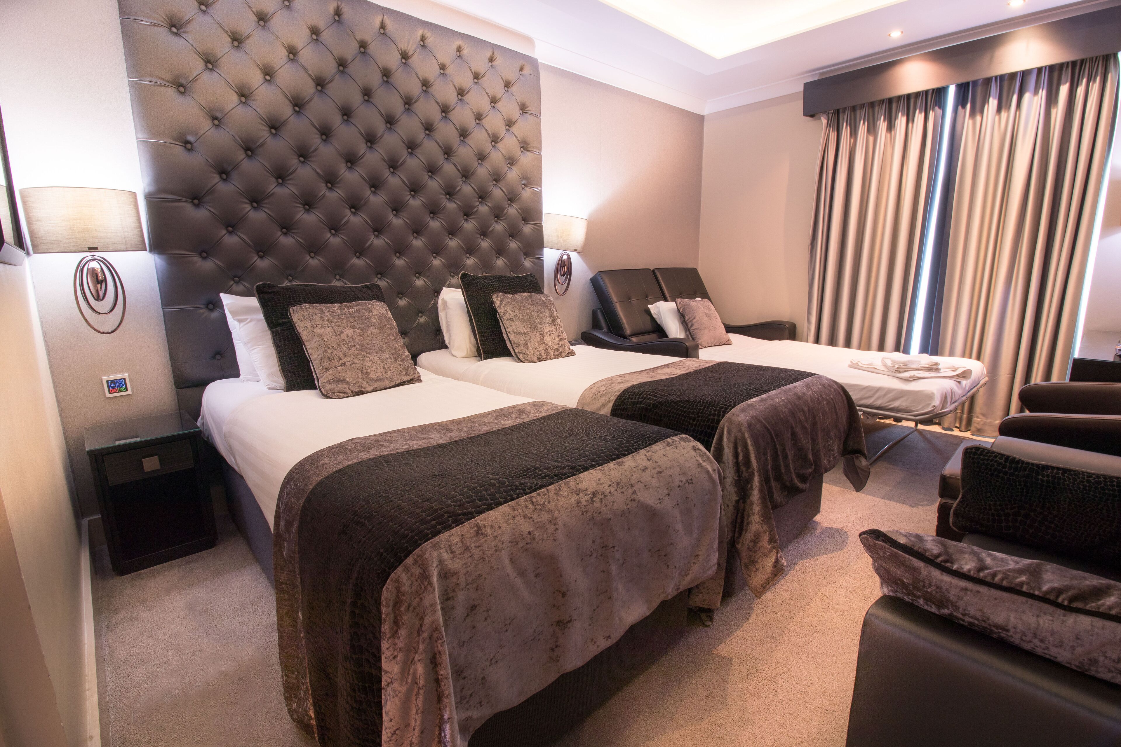 A hotel room with two beds, a tufted headboard, a massage chair, and a small table with a lamp.