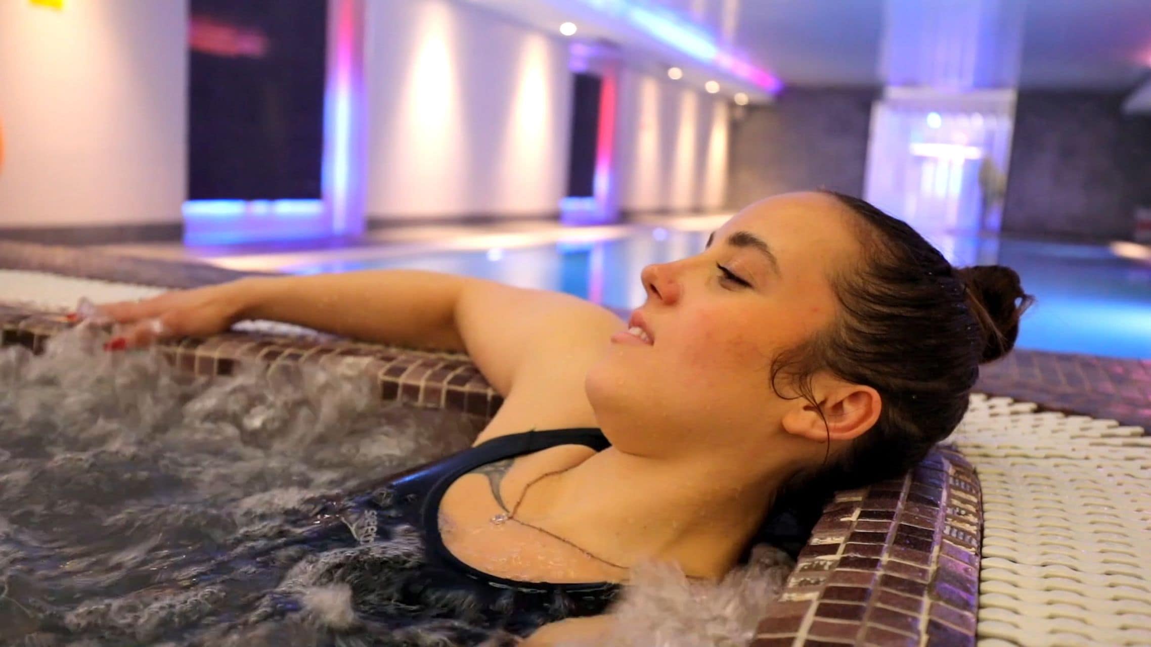 A person relaxing in a hot tub with a tiled floor and a pool in the background.
