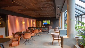 A modern indoor space with a long counter, multiple tables and chairs, and a large screen displaying a person. - Grand Hotel Struer (Struer)