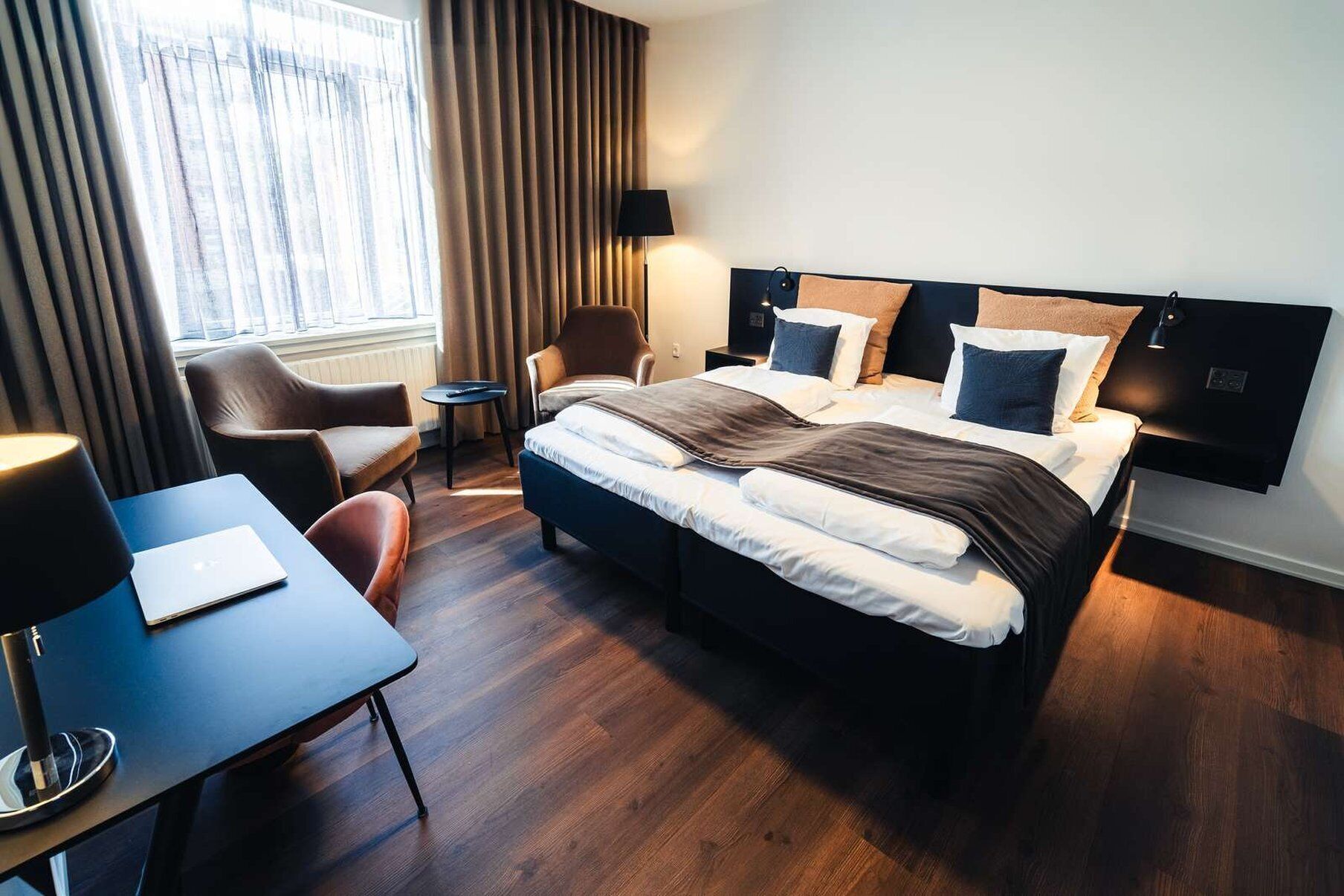 A modern hotel room with a large bed, a desk with a laptop, a chair, and a window with curtains.